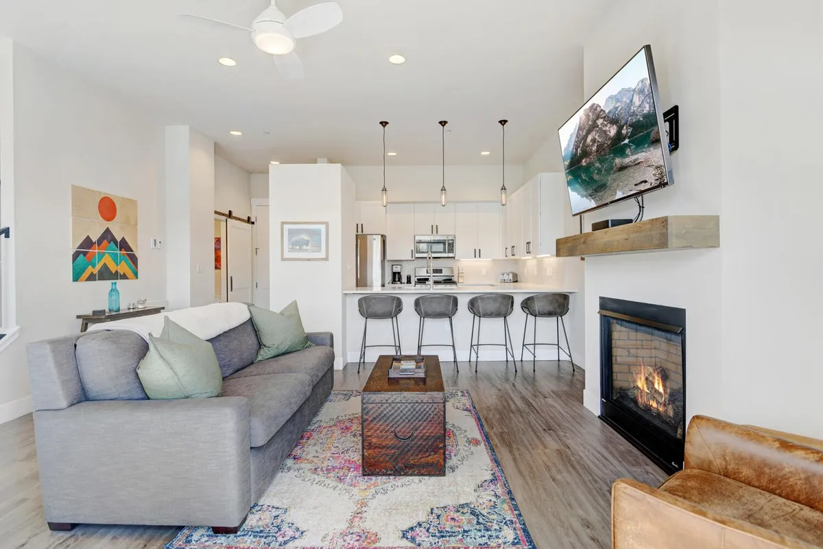 The image showcases a stylish, open-concept living area with a plush sofa and coffee table centered around a glowing fireplace. The adjacent modern kitchen island hints at easy group meals, while the overall ambiance is bright, cozy, and welcoming—ideal for socializing, relaxing, and enjoying mountain mornings or fireside evenings.