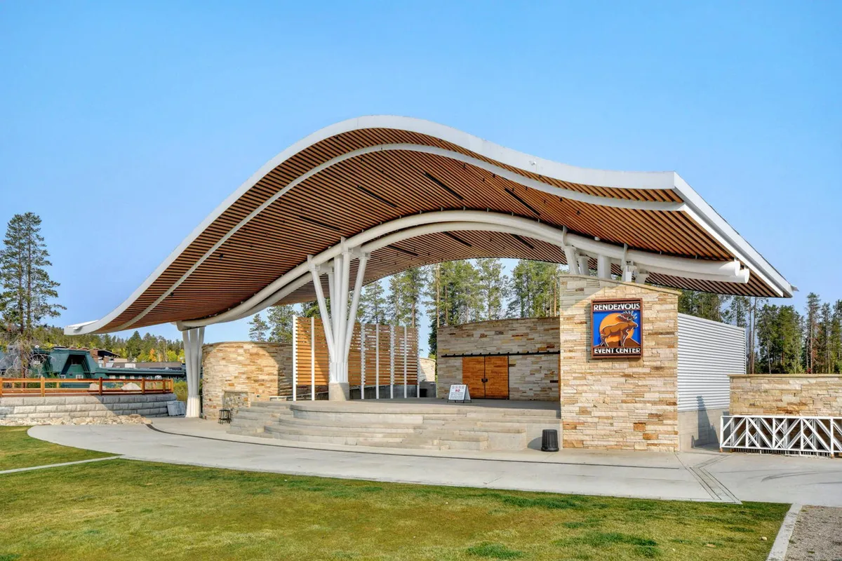 This exterior image spotlights the iconic curved roof structure, stone façade, and open stage area at Hideaway Park, an inviting outdoor venue known for hosting community events, concerts, and family-friendly gatherings. The open space suggests opportunities for picnics, play, and enjoying the summer sun.
