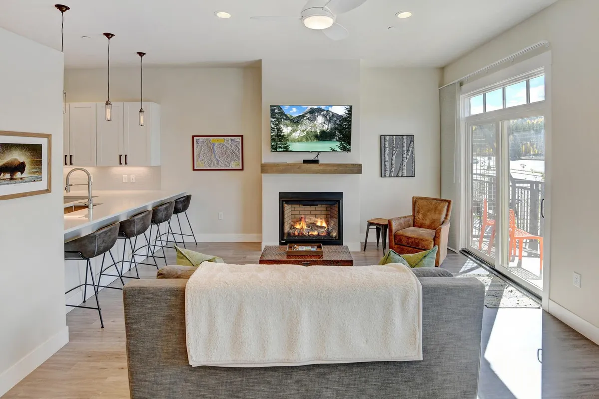 A stylish, open interior featuring a glowing fireplace, plush sofa, television, bar counter, and expansive windows offering a glimpse of the outdoors. The ambiance is both warm and modern, ideal for enjoying sunsets, socializing, or a peaceful evening in.
