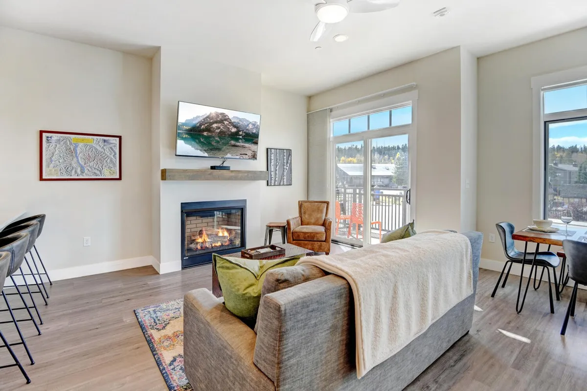 A spacious, open-concept living area featuring a cozy sofa facing a gas fireplace and a large wall-mounted TV. The room is bathed in natural light from expansive windows, with a dining table visible nearby—showcasing the seamless flow between living, dining, and kitchen spaces.