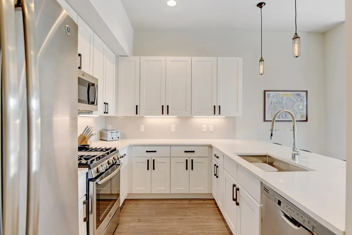 A bright, modern kitchen featuring stainless steel appliances, white cabinetry, quartz countertops, and pendant lighting. The space feels open and functional—ideal for meal prep or informal gatherings, with framed artwork adding a touch of personality.