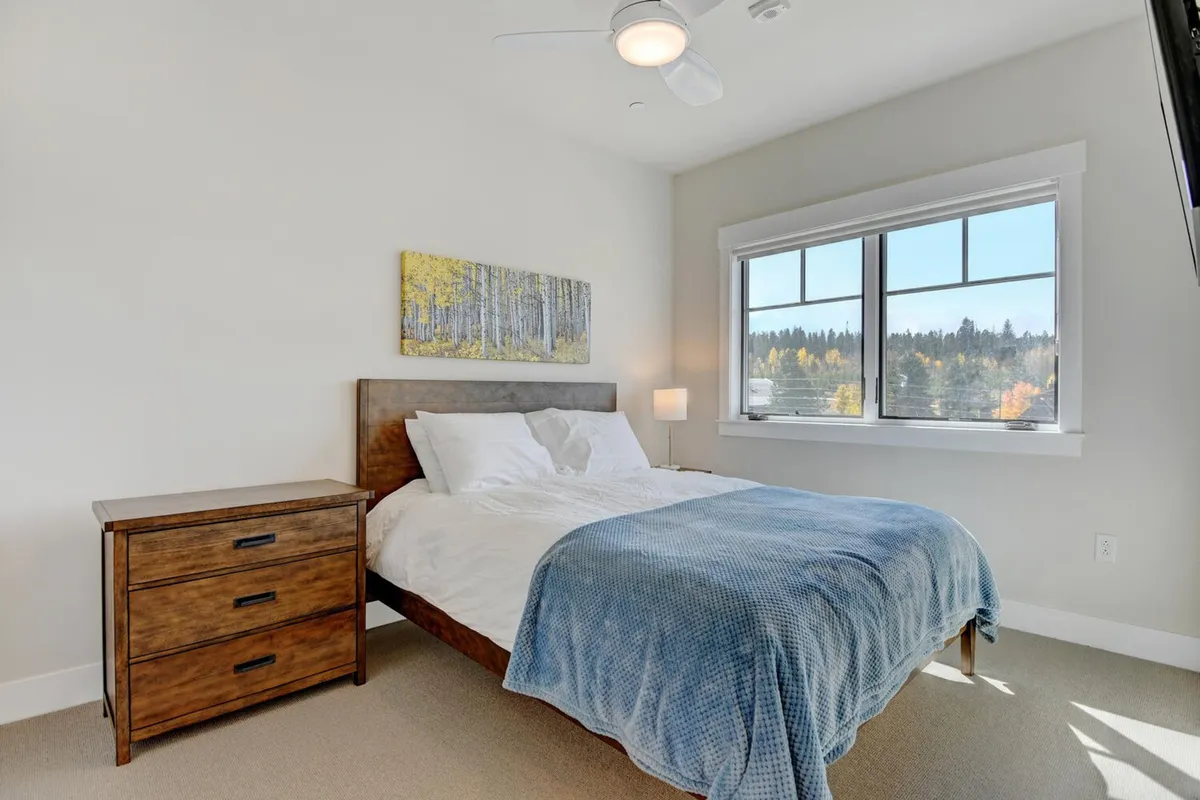 A tranquil bedroom featuring a plush bed with soft linens, a nightstand with a reading lamp, tasteful artwork on the wall, and a sunlit window that offers a soothing view of the outdoors. The setting is cozy and welcoming, ideal for restful nights.