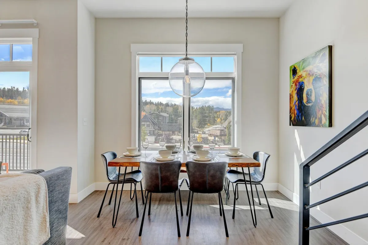 A bright dining area with a large table set near expansive windows, providing natural light and scenic views. Artwork featuring a bear adds a touch of character, and the space is arranged for group meals and gatherings.